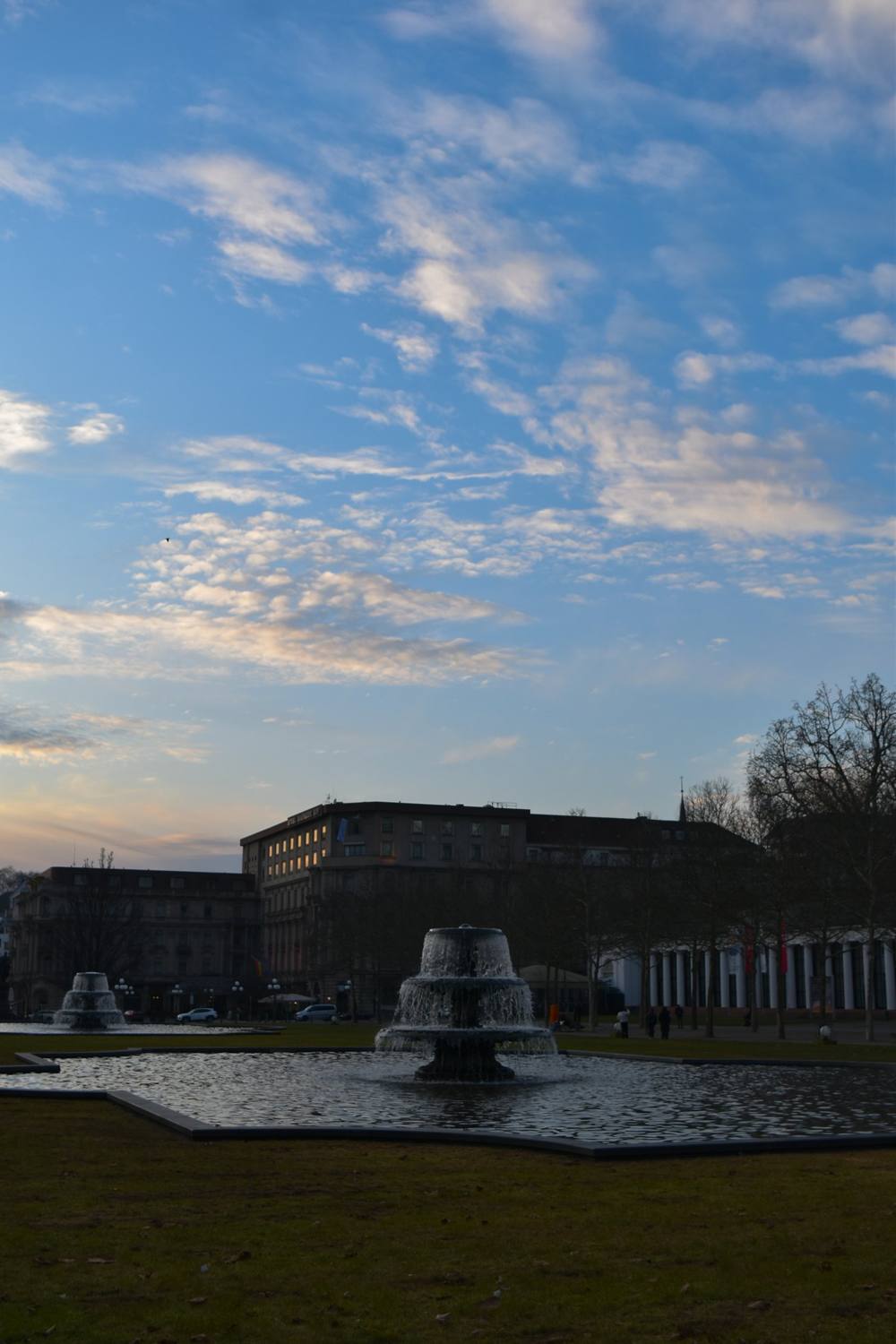 Fountain on a lawn at dusk, buildings and a row of columns behind
